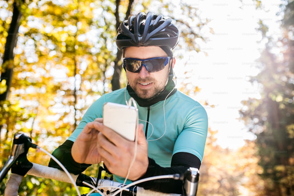 Joven deportista guapo montando su bicicleta al aire libre en la naturaleza soleada del otoño. Sosteniendo el teléfono inteligente, escuchando música.