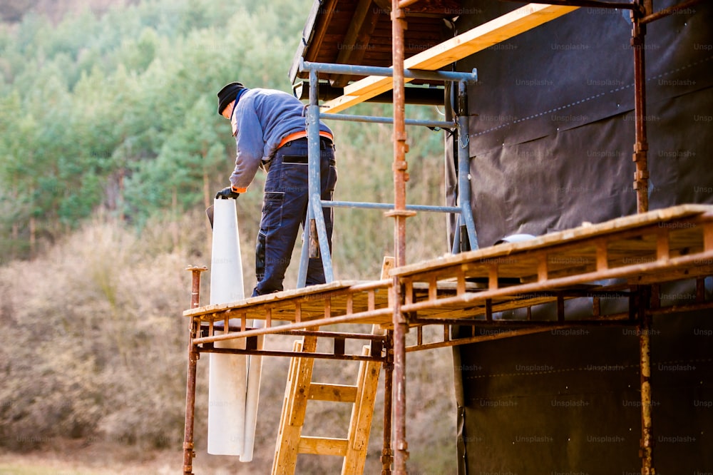 Trabalhador da construção civil em pé na fachada da casa com isolamento térmico de andaime, colocando papel alumínio preto