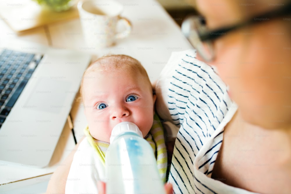 Close up of unrecognizable mother holding her baby son in the arms, feeding him with milk from bottle, laptop and coffee cup on laid on table