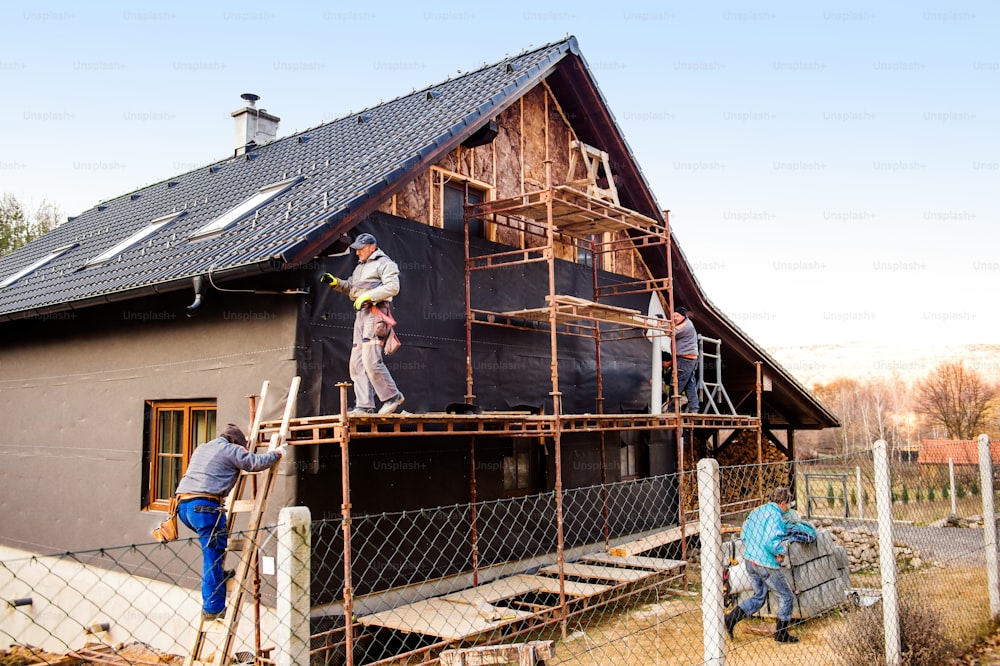 Des ouvriers de la construction debout sur un échafaudage isolant thermiquement la façade de la maison avec de la laine de verre et du papier d’aluminium noir.