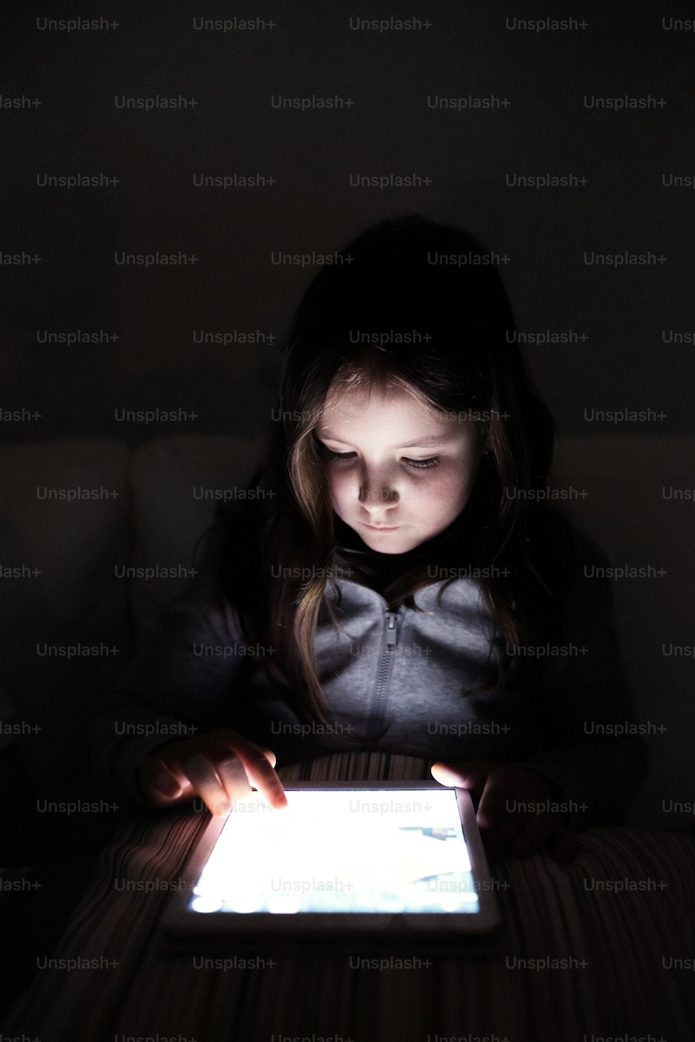 Little girl playing with tablet, sitting on sofa in dark room at night.