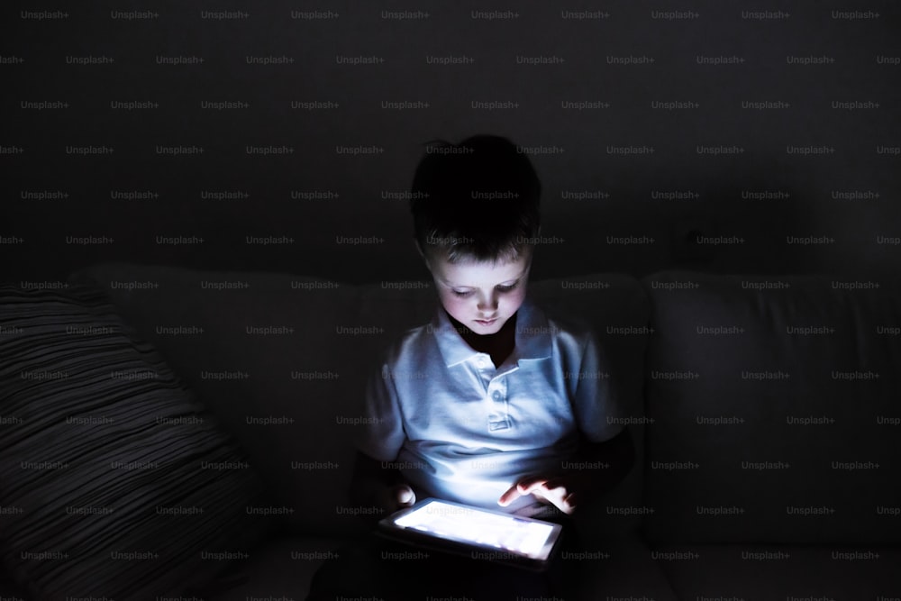 Little boy playing with tablet, sitting on sofa in dark room at night.