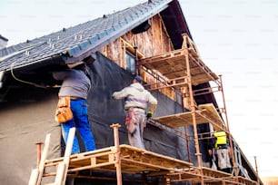 Construction workers standing on scaffold thermally insulating house facade with glass wool and black foil.