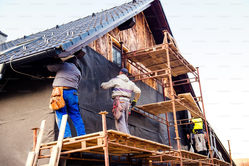 Construction workers standing on scaffold thermally insulating house facade with glass wool and black foil.