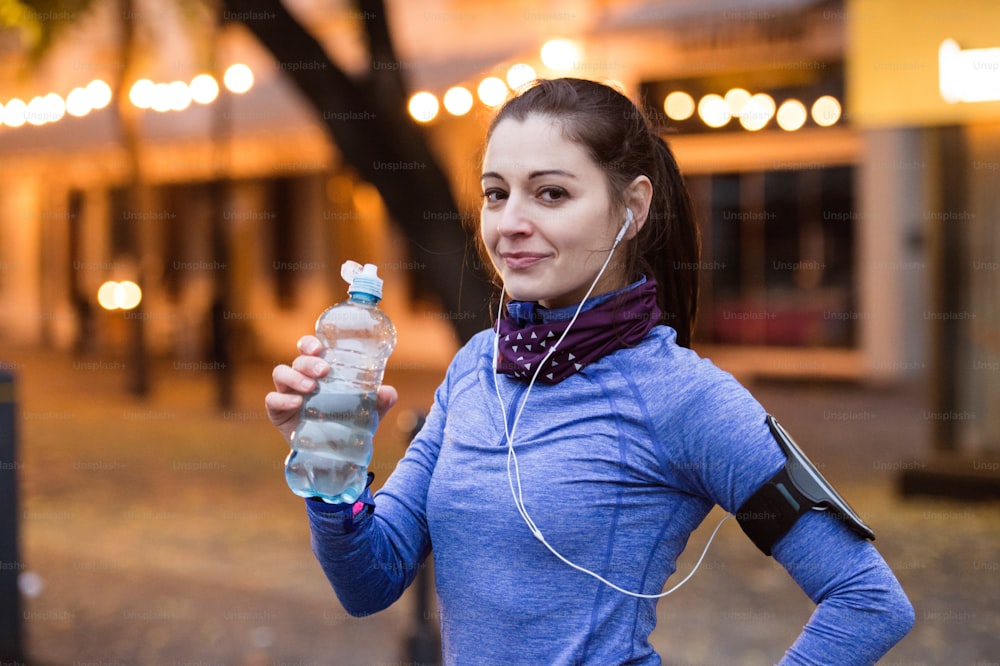 Beautiful young woman with smart phone and earphones, listening music, resting, drinking water from bottle. Running in the illuminated night town.