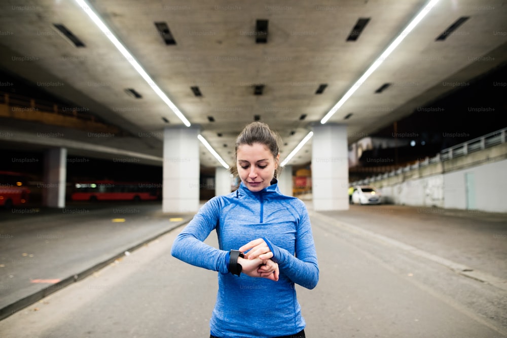 Bela jovem mulher sob a ponte na cidade à noite com smartwatch, usando um aplicativo de fitness para acompanhar o progresso da perda de peso, meta de corrida ou resumo de sua corrida.