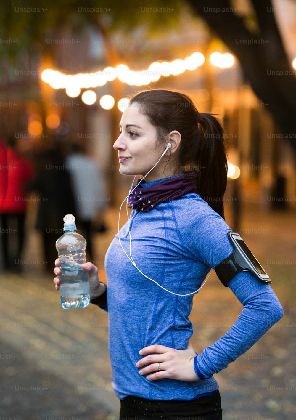 Bella giovane donna con smart phone e auricolari, ascoltando musica, riposando, bevendo acqua dalla bottiglia. Correre nella città notturna illuminata.