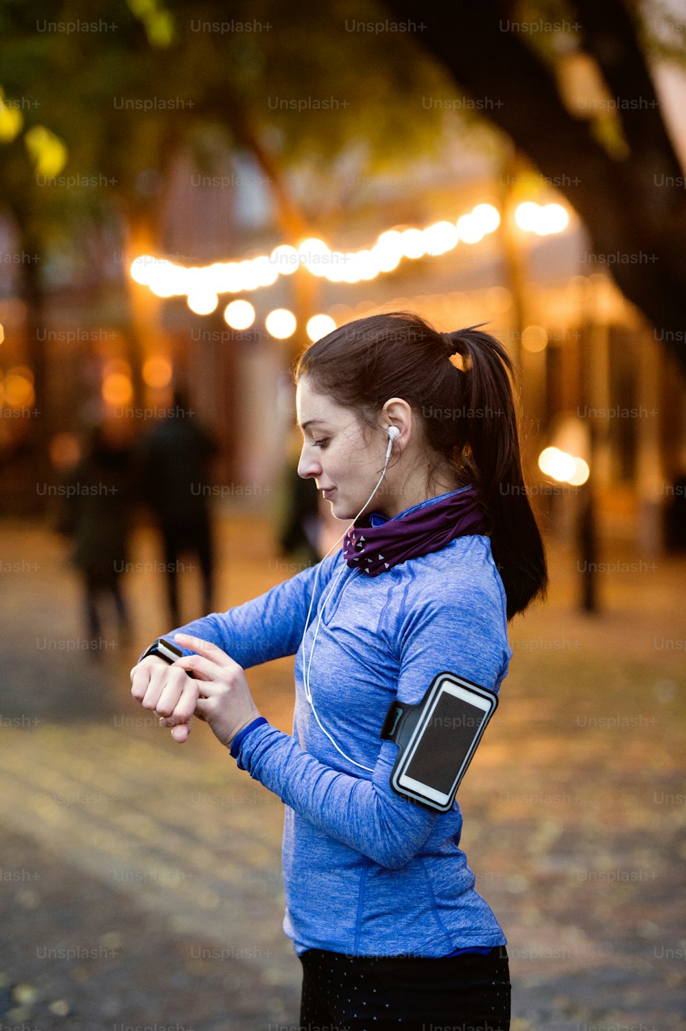 Beautiful young woman in the town in the evening with smart phone, smartwatch and earphones, listening music. Using a fitness app for tracking weight loss progress, running goal or summary of her run.