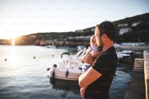 Beau jeune homme debout sur une jetée en bois tenant son bébé dans ses bras profitant de leur temps au bord de la mer.