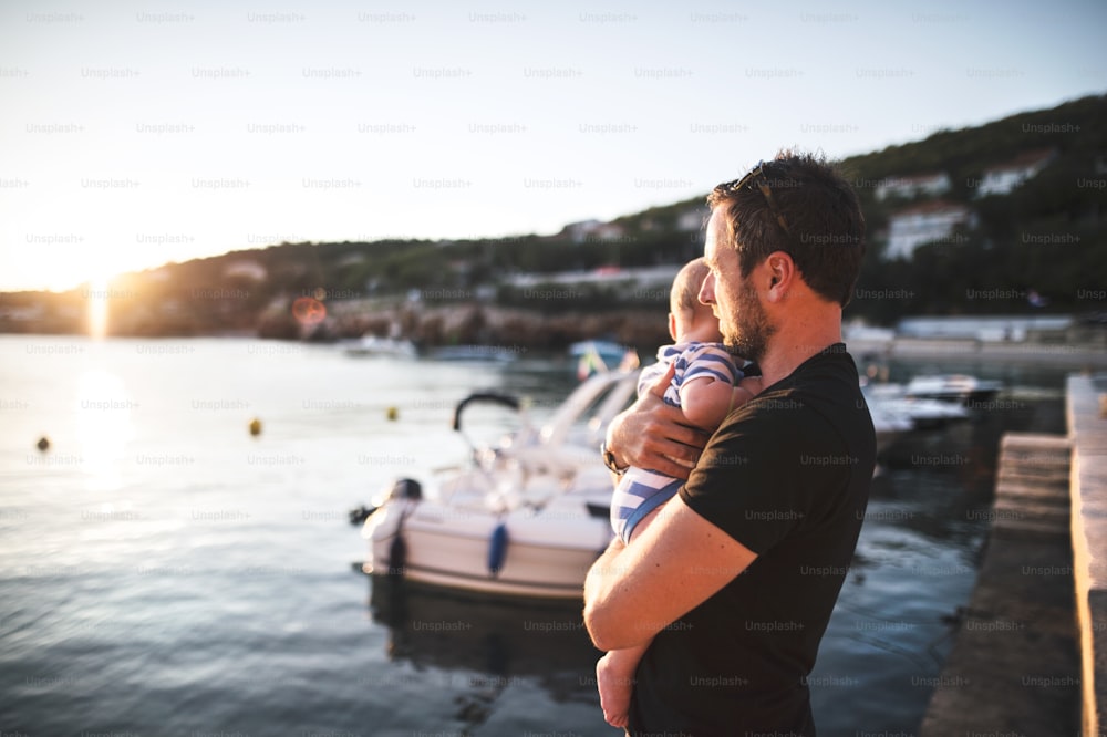Hübscher junger Mann, der auf einem hölzernen Pier steht und seinen kleinen Sohn in den Armen hält und ihre Zeit am Meer genießt.