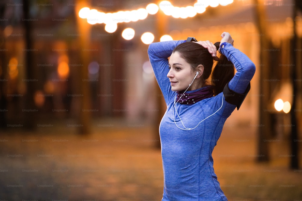 Beautiful young runner with armband and earphones, listening music, warming up and stretching in the illuminated night town.