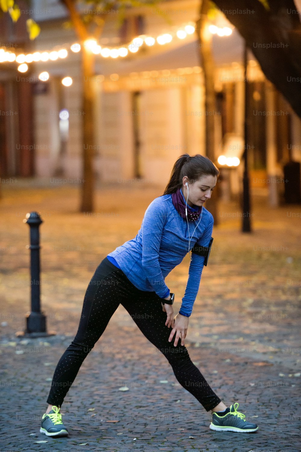Beautiful young runner with armband and earphones, listening music, warming up and stretching legs in the illuminated night town.