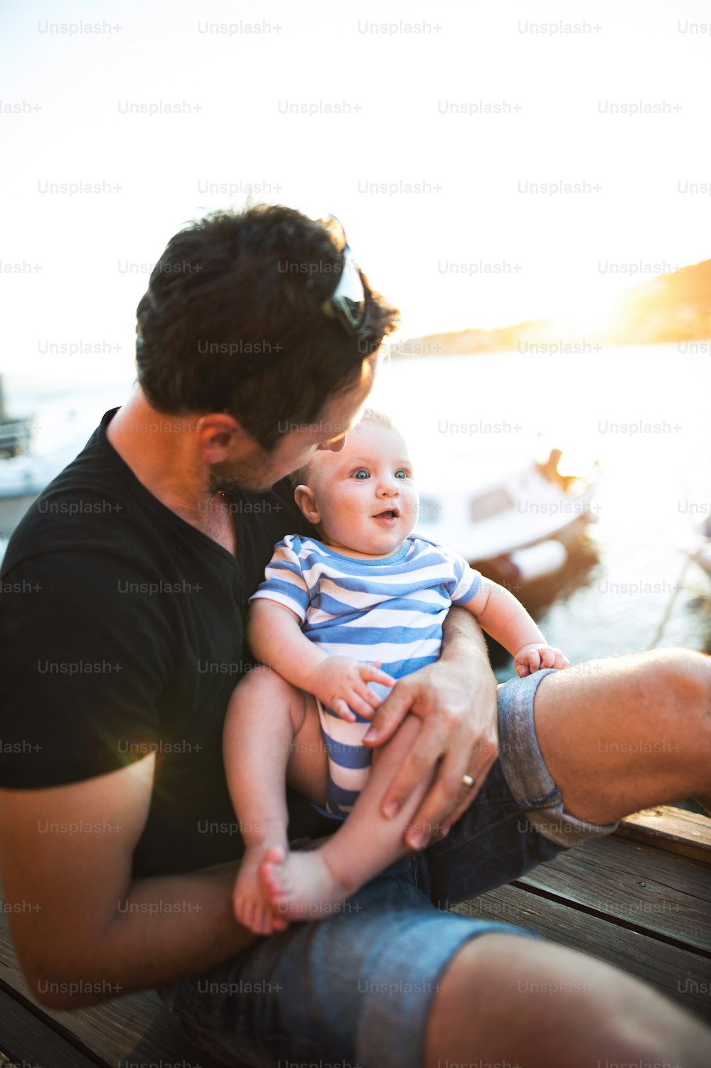 Handsome young man sitting on wooden pier holding his baby son in his arms enjoying their time at seaside.