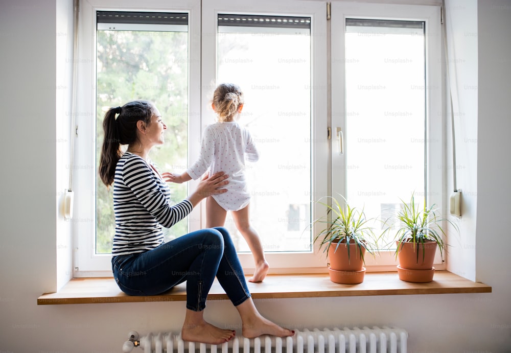 Beautiful young mother sitting on window sill with her cute little daughter looking out of window