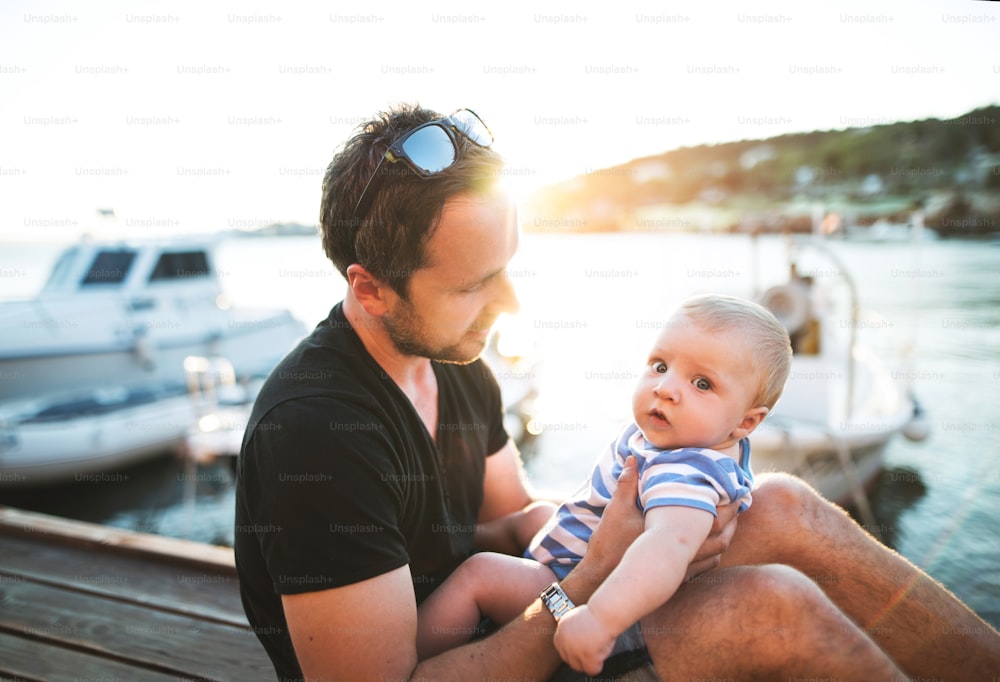 Handsome young man sitting on wooden pier holding his baby son in his arms enjoying their time at seaside.
