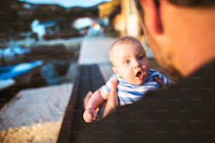 Unrecognizable young man sitting on wooden pier holding baby son in his arms enjoying their time at seaside. Little boy wondering with mouth and eyes open wide.