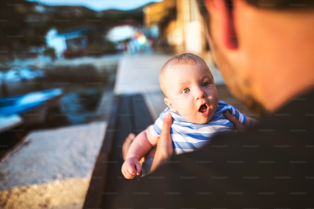 Unrecognizable young man sitting on wooden pier holding baby son in his arms enjoying their time at seaside. Little boy wondering with mouth and eyes open wide.