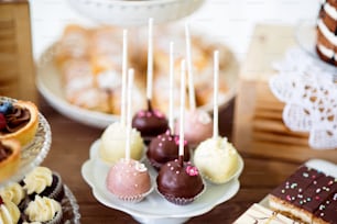 Table avec cake pops blancs, roses et chocolat sur une assiette, tartes et cupcakes sur cakestand. Barre chocolatée.