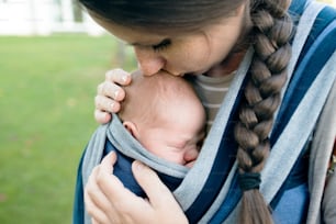 Beautiful young mother kissing her newborn baby son in sling outside in green nature.