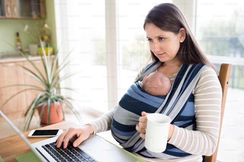 Schöne junge Mutter mit ihrem neugeborenen Sohn im Tragetuch zu Hause, am Tisch sitzend, auf der Notebook-Tastatur schreibend, eine Tasse Tee in der Hand halten