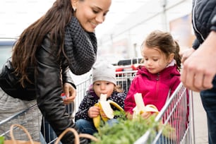 Young parents unloading groceries from shopping cart to the back of the car. Two little daughters sitting in a trolley, eating babanas. Autumn rainy day.