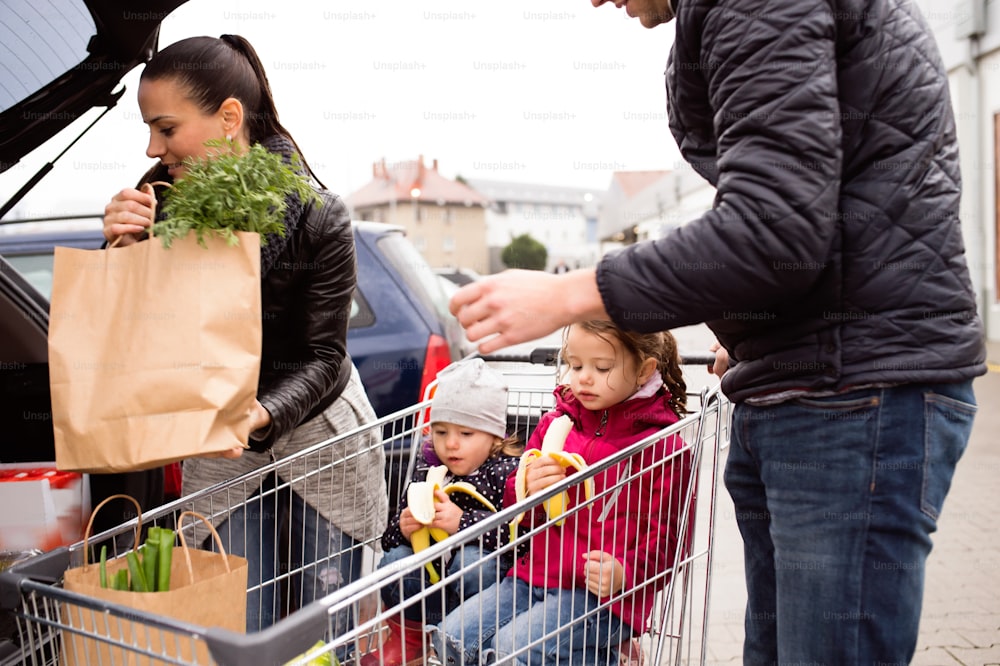 Young parents unloading groceries from shopping cart to the back of the car. Two little daughters sitting in a trolley, eating babanas. Autumn rainy day.