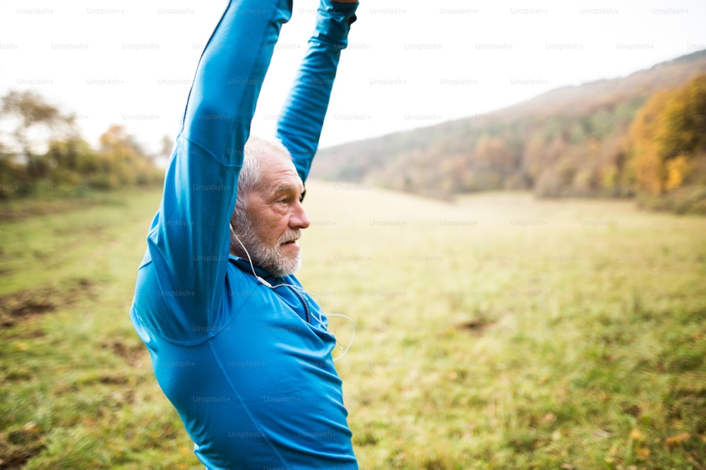 Senior runner in sunny autumn nature with earphones, listening music, doing stretching.