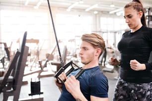 Young handsome fit man with his personal trainer working out on pull-down machine in gym. Bodybuilder exercising with cable weight machine.