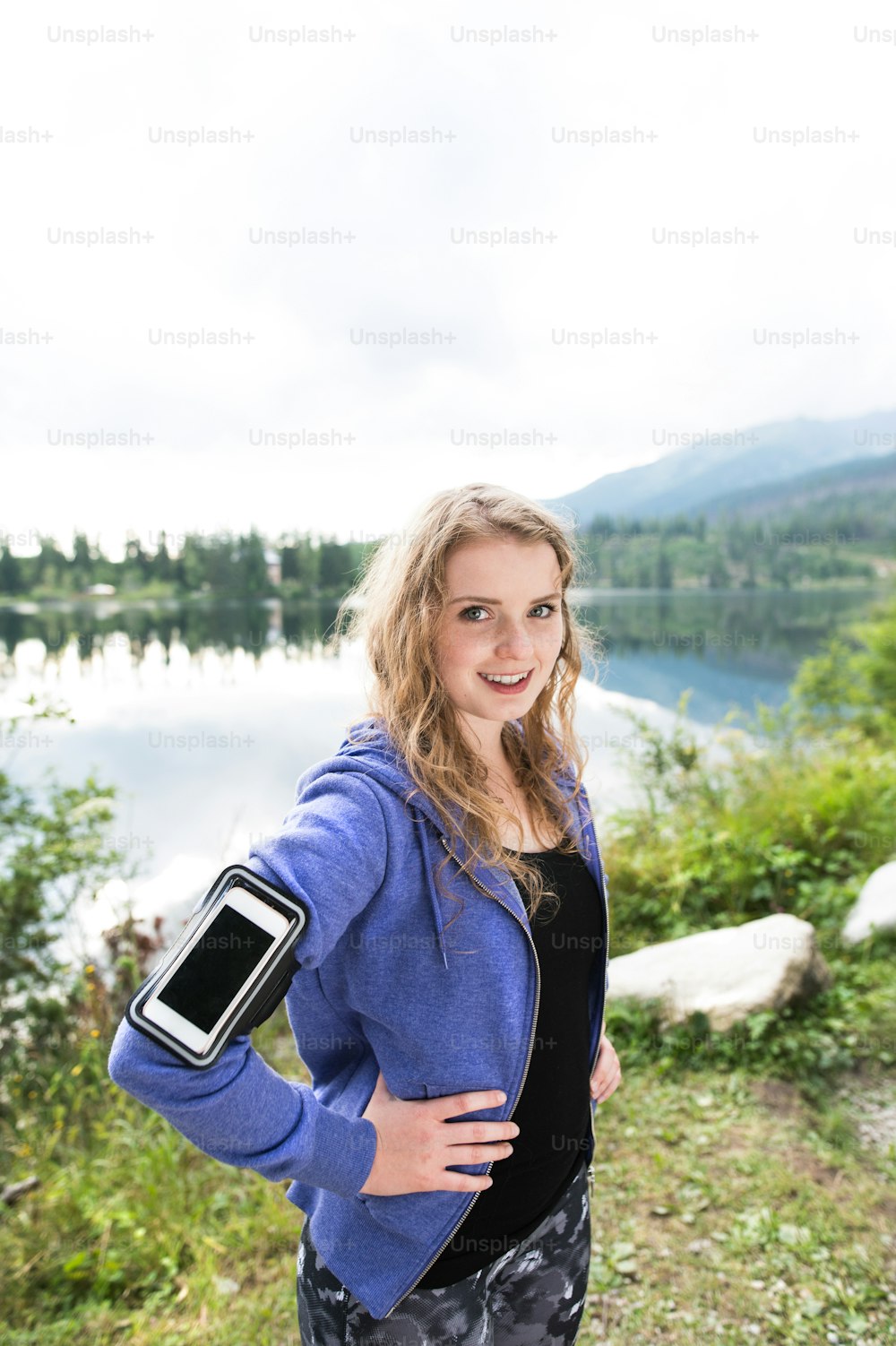 Beautiful young woman with smart phone in blue sweatshirt at the lake in green nature. Using a fitness app for tracking weight loss progress, running goal or summary of her run.