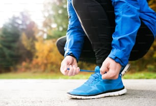 Unrecognizable runner in blue jacket outside in colorful sunny autumn nature, tying shoelaces