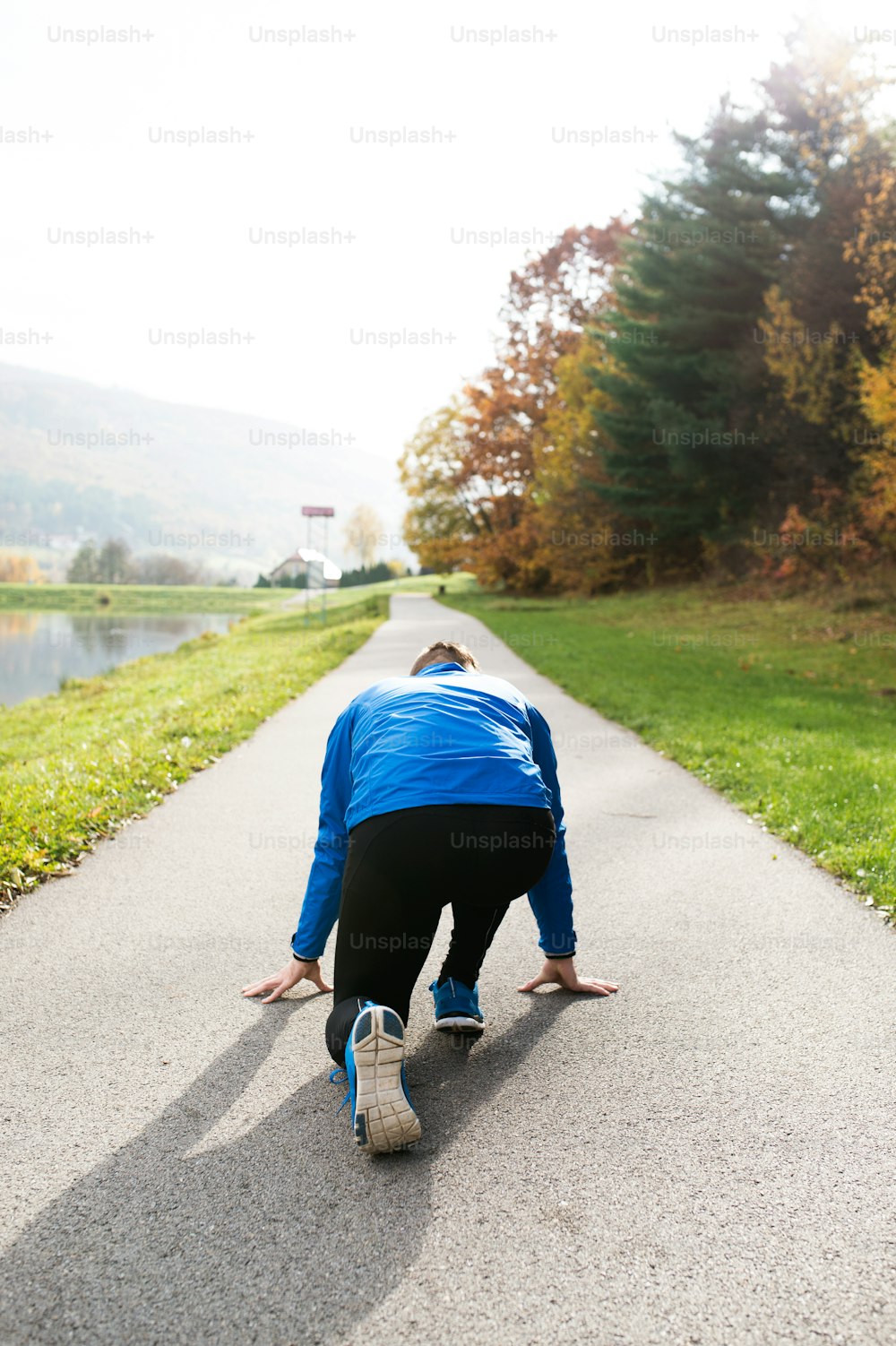 Young runner in blue jacket on an asphalt path leading through green grass in steady position. Trail runner training for cross country running outside in colorful sunny autumn nature.
