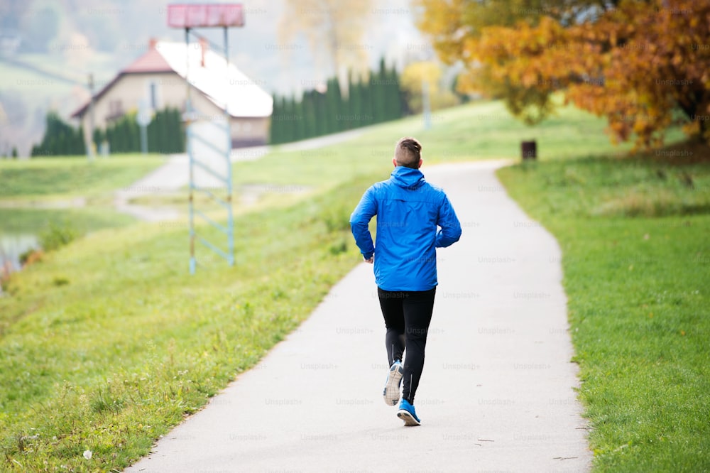Young athlete in blue jacket running outside in colorful sunny autumn nature on an asphalt path leading through green grass. Trail runner training for cross country running.
