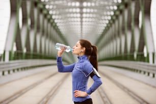 Beautiful young woman with smart phone and earphones, listening music, running in the city on green steel bridge, resting, drinking water from bottle.