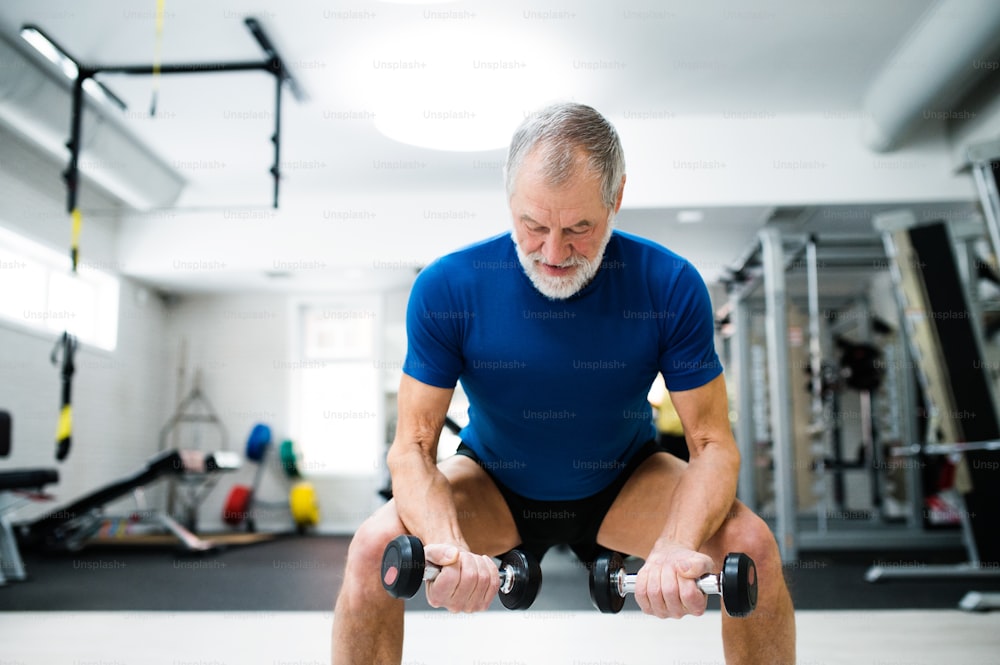 Senior Mann in Sportkleidung im Fitnessstudio trainiert mit Gewichten und hockt gleichzeitig.