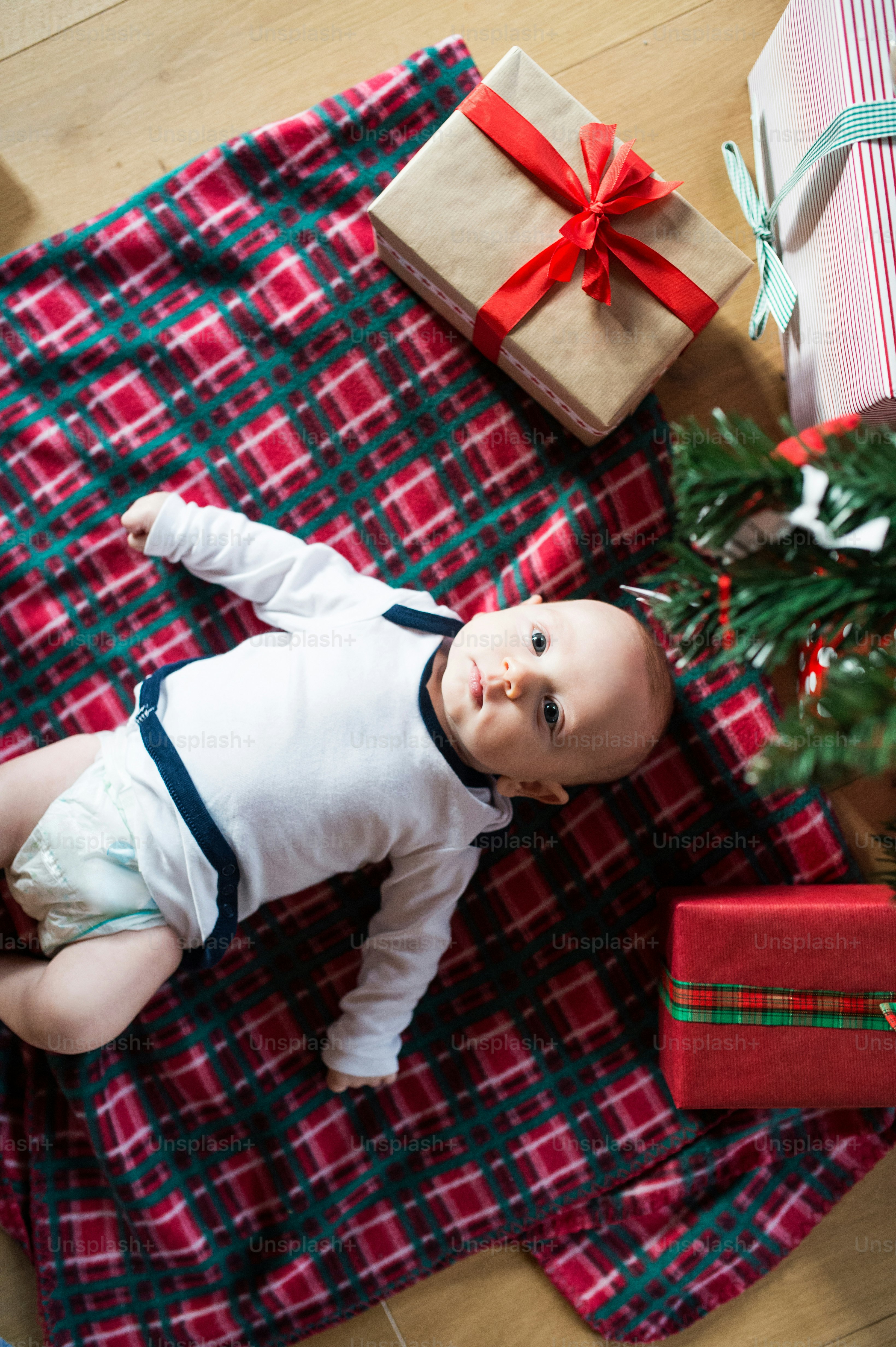 Cute little baby boy under Christmas tree lying among presents on checked blanket