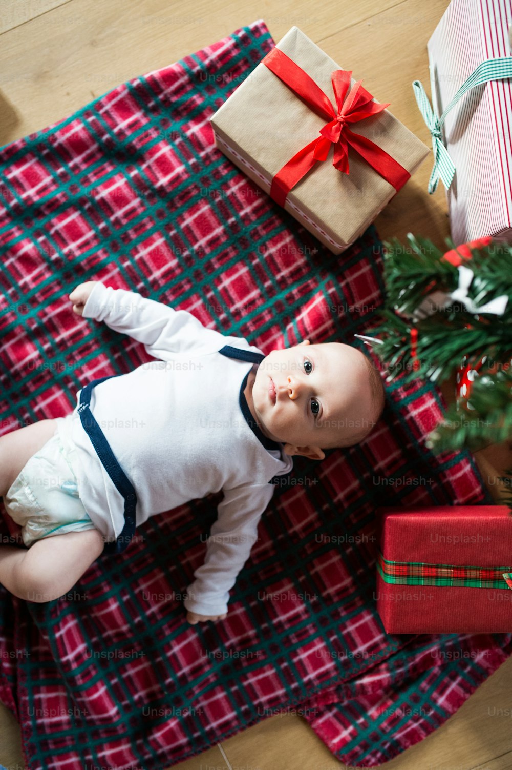 Cute little baby boy under Christmas tree lying among presents on checked blanket.
