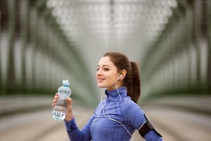Hermosa mujer joven con teléfono inteligente y auriculares, escuchando música, corriendo en la ciudad en un puente de acero verde, descansando, bebiendo agua de la botella.
