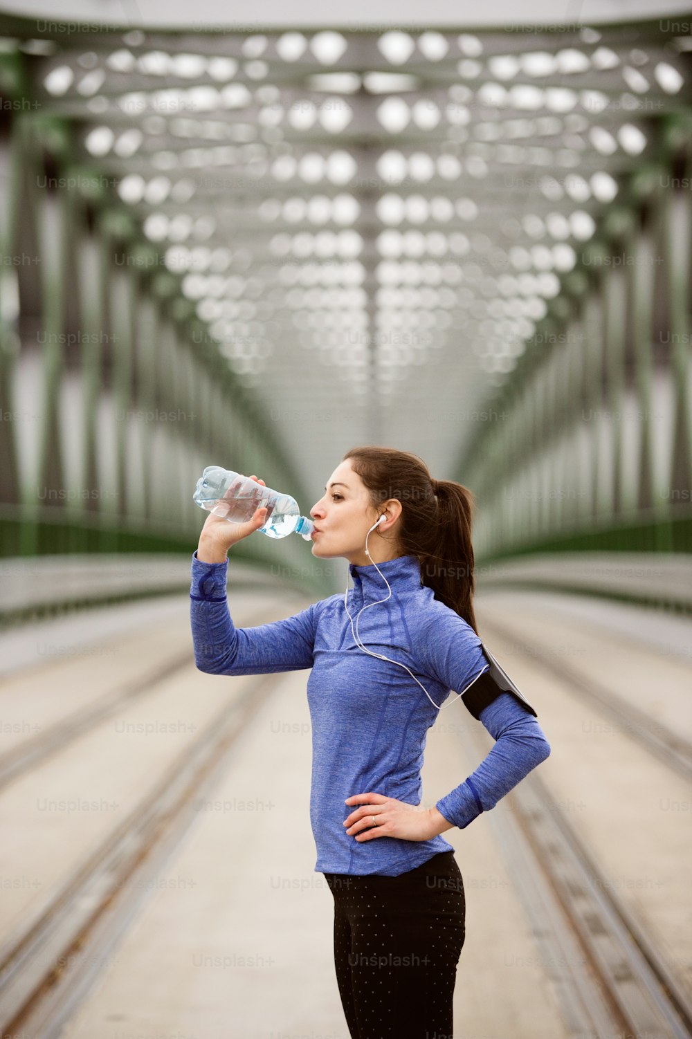 Beautiful young woman with smart phone and earphones, listening music, running in the city on green steel bridge, resting, drinking water from bottle.