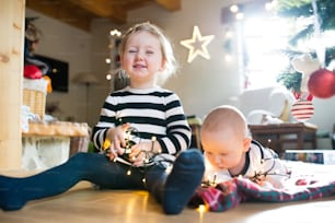 Süßes kleines Mädchen mit seinem kleinen Bruder unter dem Weihnachtsbaum, das auf einer karierten Decke liegt, die sich in einer Lichterkette verfängt.