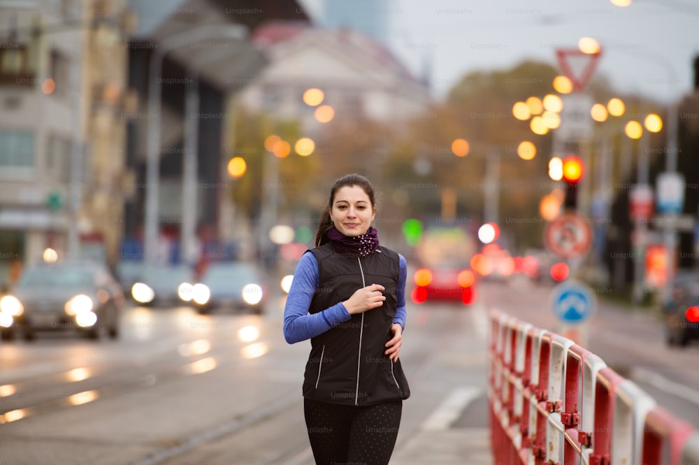 Beautiful young woman in black vest running in the city against road with cars