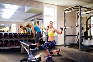 Close up of two attractive fit women in gym exercising with weights and bar while doing lunges