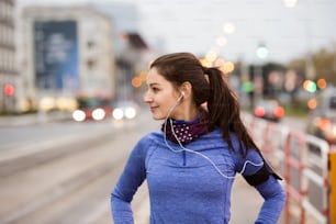 Hermosa mujer joven con teléfono inteligente y auriculares, escuchando música, corriendo en la ciudad contra la carretera con automóviles