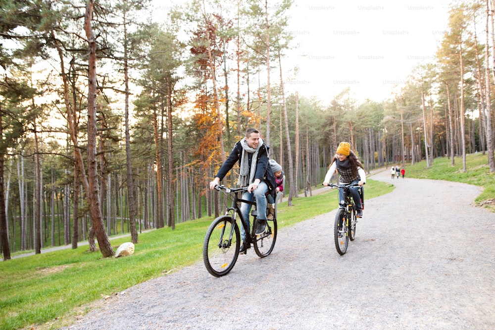 Bella giovane famiglia con due figlie in seggiolini per biciclette in vestiti caldi che pedalano all'aperto nella natura autunnale