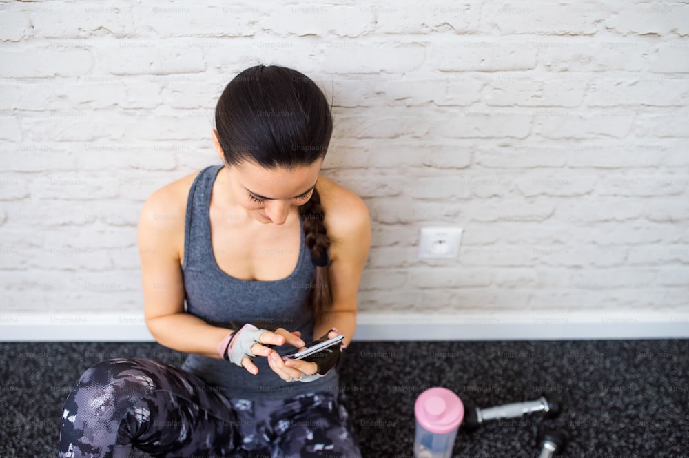 Smiling attractive fit woman in gym sitting on a floor holding smart phone against white brick wall, water bottle, weights
