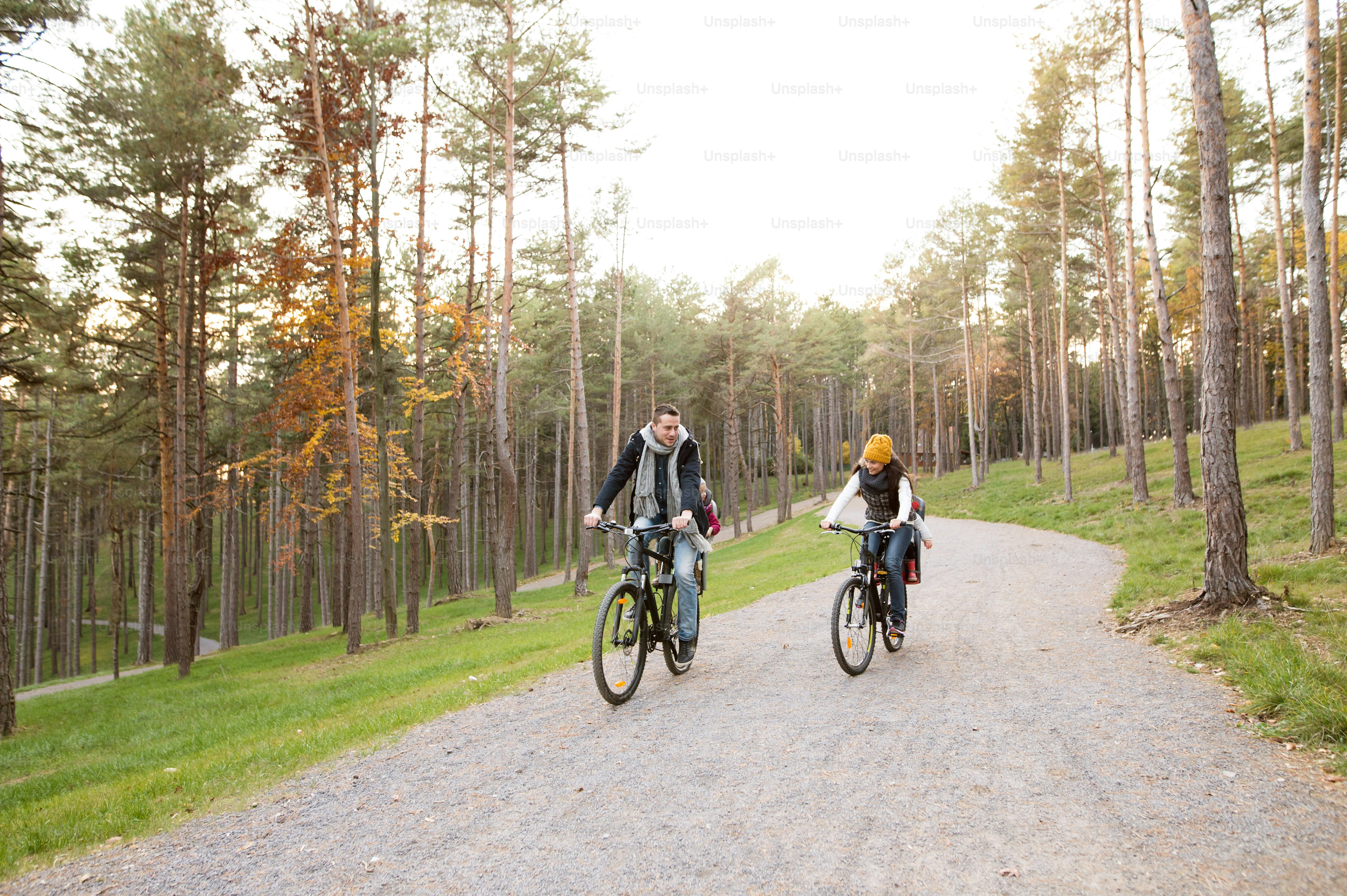 Schöne junge Familie mit zwei Töchtern in Fahrradsitzen in warmen Kleidern, die draußen in der Herbstnatur radeln