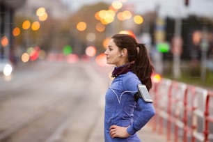 Beautiful young woman with smart phone and earphones, listening music, running in the city against road with cars