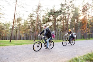 Bella giovane famiglia con due figlie in seggiolini per biciclette in vestiti caldi che pedalano all'aperto nella natura autunnale