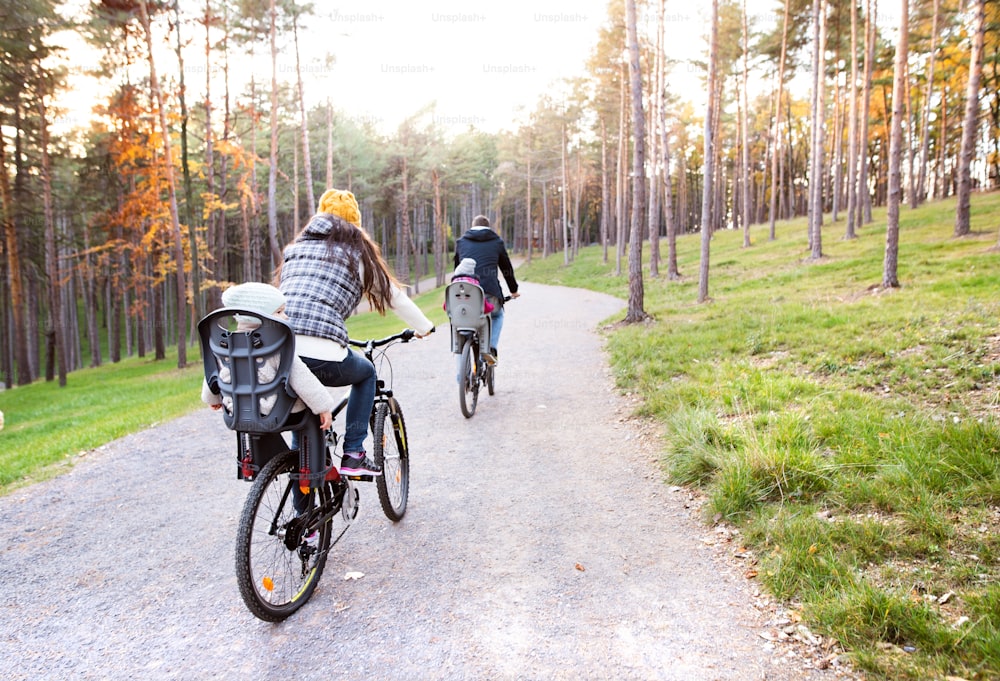 Hermosa familia joven con dos hijas en asientos de bicicleta con ropa de abrigo que andan en bicicleta al aire libre en la naturaleza otoñal. Vista trasera.