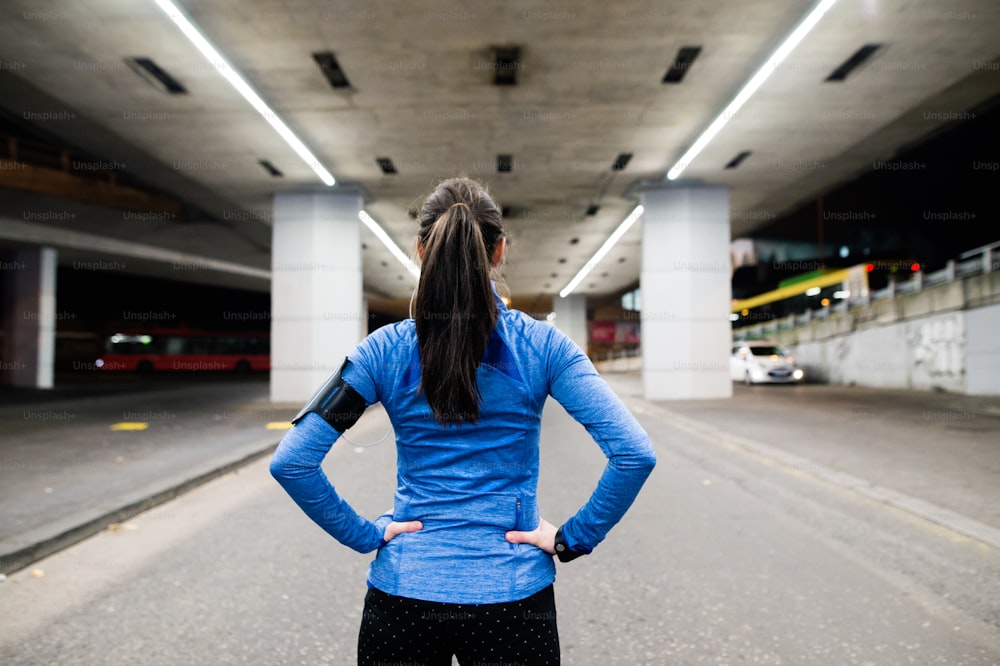 Beautiful young runner under the bridge in the town in the evening resting, arms on hips. Rear view.