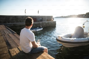Hübscher junger Mann, der die Zeit am Meer genießt, auf einem hölzernen Pier sitzt und ein Glas Weißwein in der Hand hält. Rückansicht.